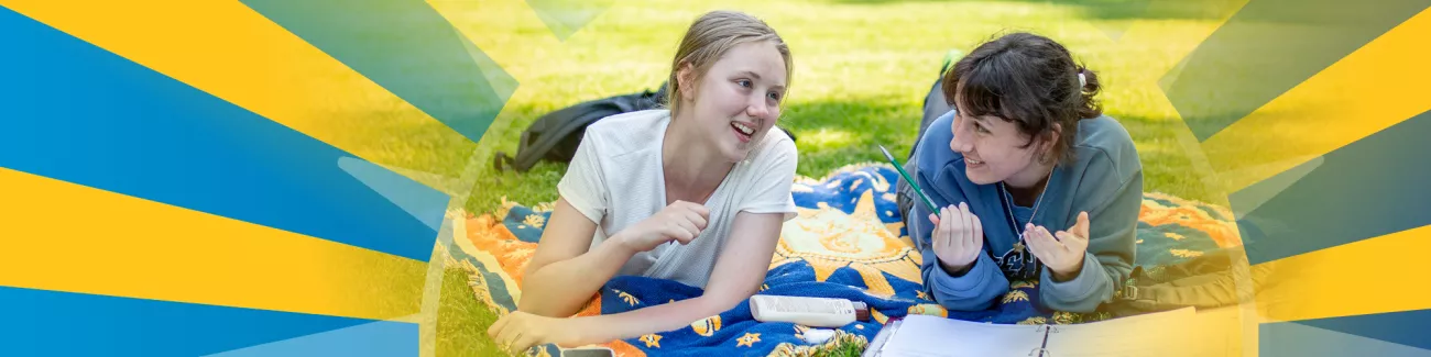 Two students lay on a blanket on a bright green lawn studying books