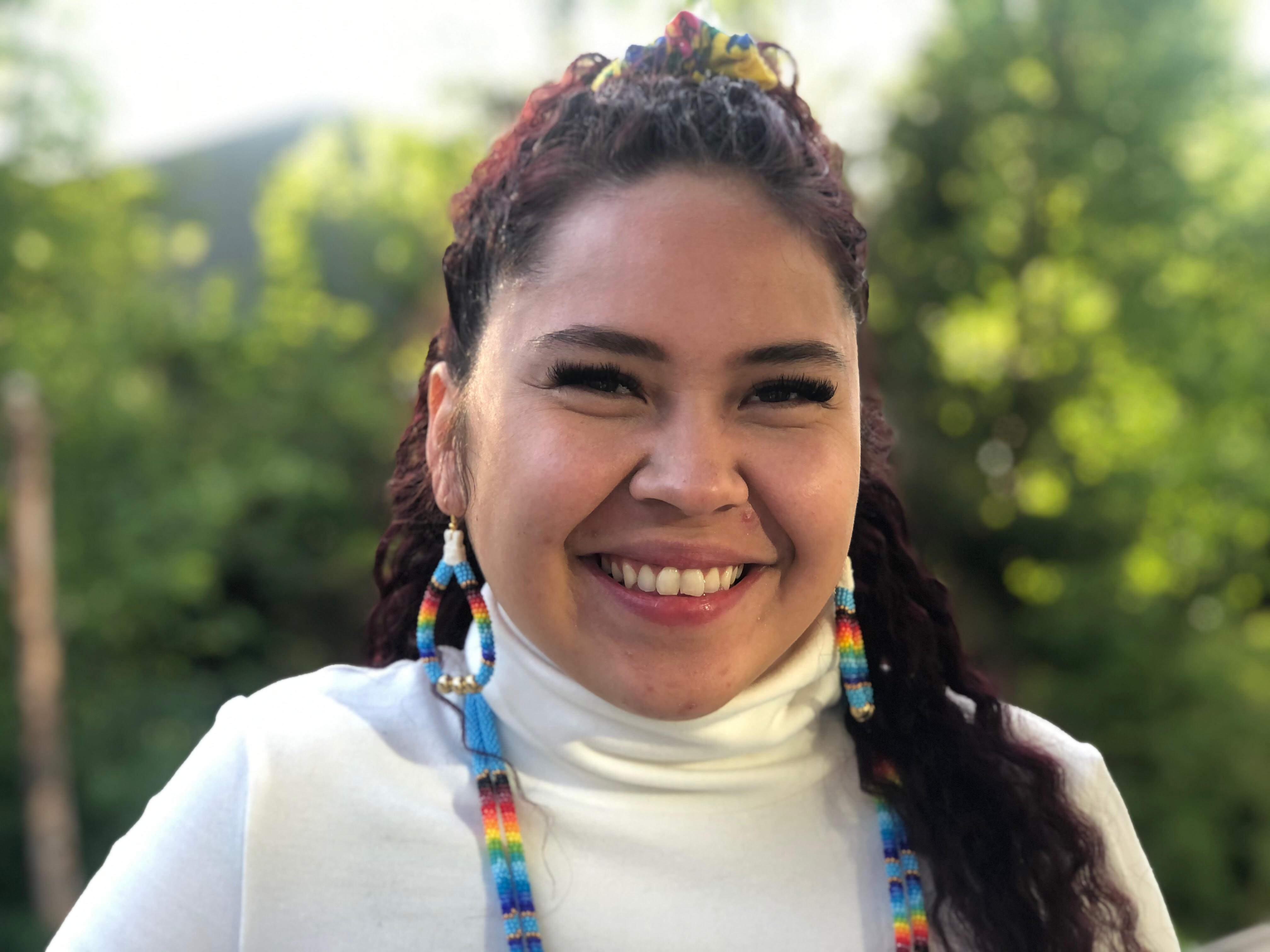 Santana, a smiling woman adorned with colorful Native American jewelry earring and matching necklace, stands outside on a sunny day.