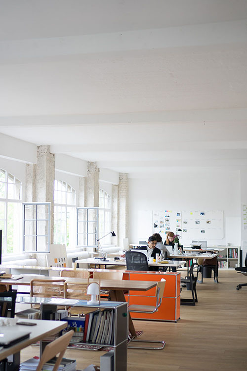 A bright white interior with a few people working at a row of rectangular tables
