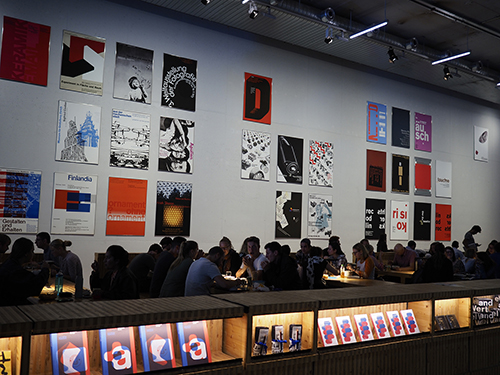 a cafeteria filled with people, with different graphic posters in a sporadic grid on the wall behind