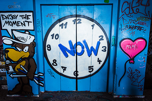 Grafitti art with a bear wearing saying 'enjoy the moment', a clock with the word 'now' int he center, and a heart balloon