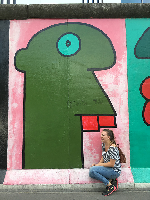 a girl sits in front of a mural of a green, abstracted cartoon man with red teeth