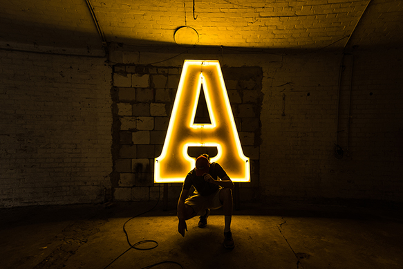 A man crouches silhouetted in front of a lighted, serif 'A'