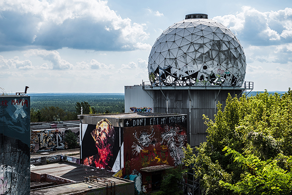 A geodesic sphere with signs of wear sits on a tower behind a square builidng with murals painted on its sides