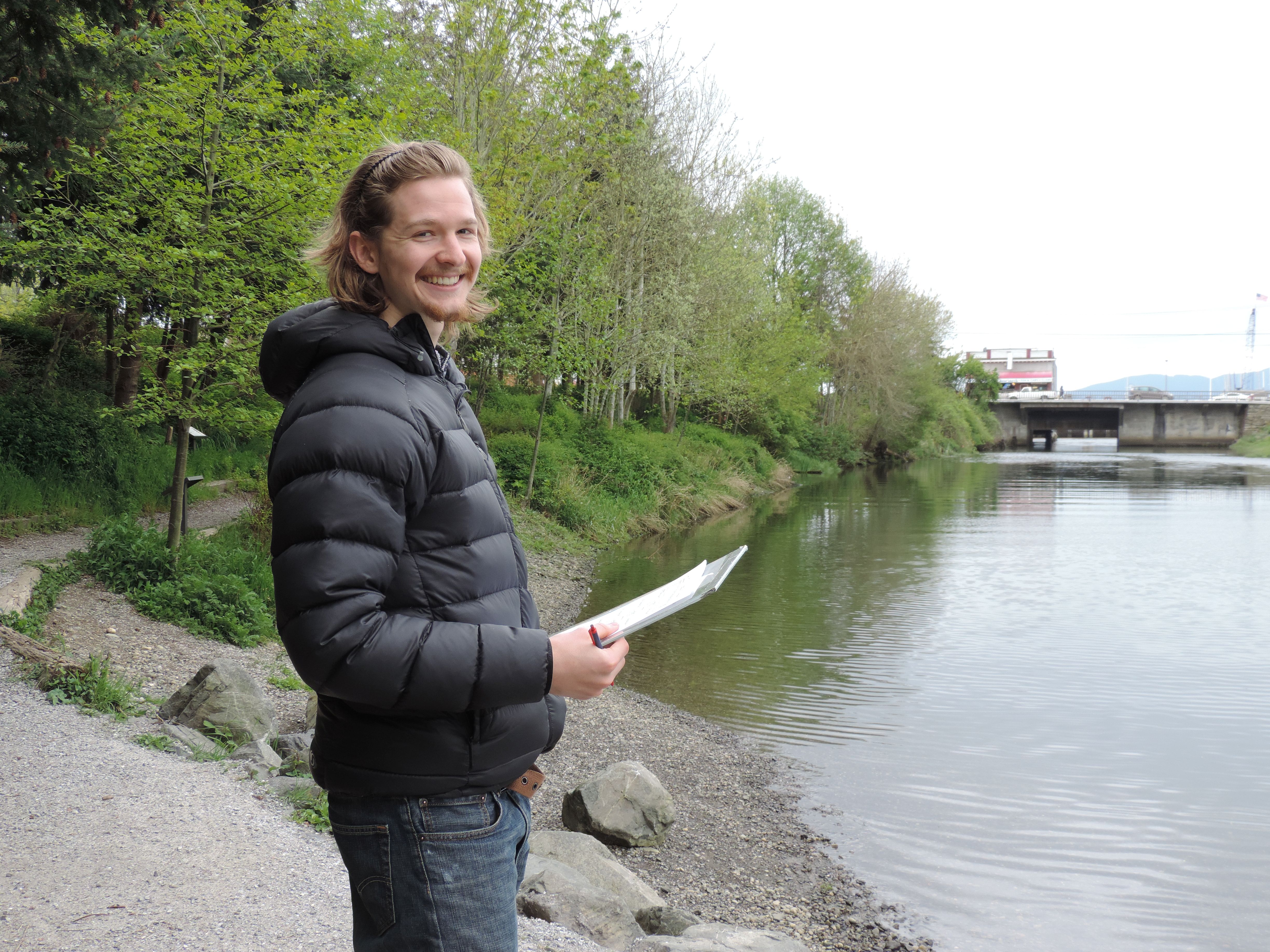 woman in a black quilted jacket holding a clipboard on a path by water