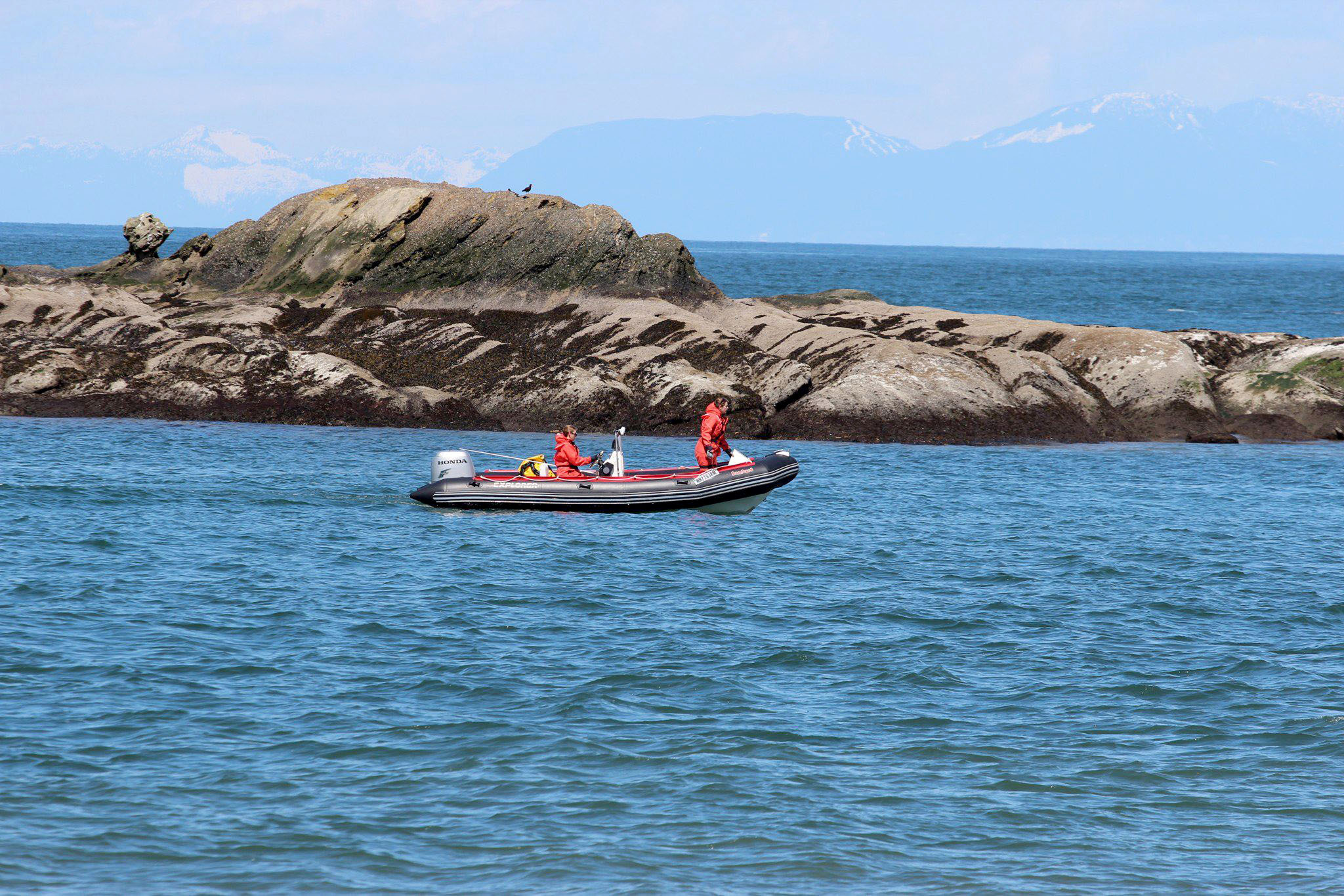 Two people in orange attire drving a boat past a rocky island.