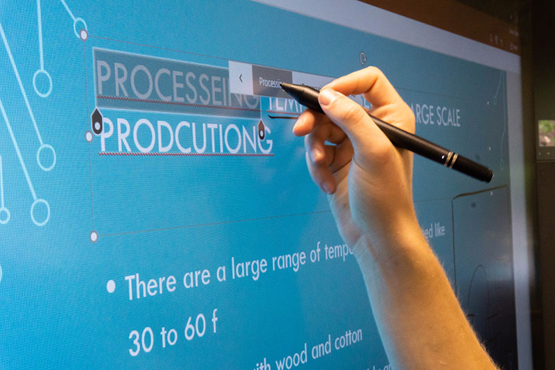 A hand holding a stylus interacting with a screen. The pen touches an onscreen 'processing' button to edit the text of a presentation slide.