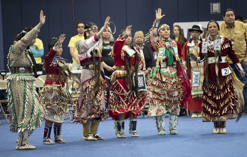 Native American tribal members across generations wear traditional clothing, with some raising their right hand