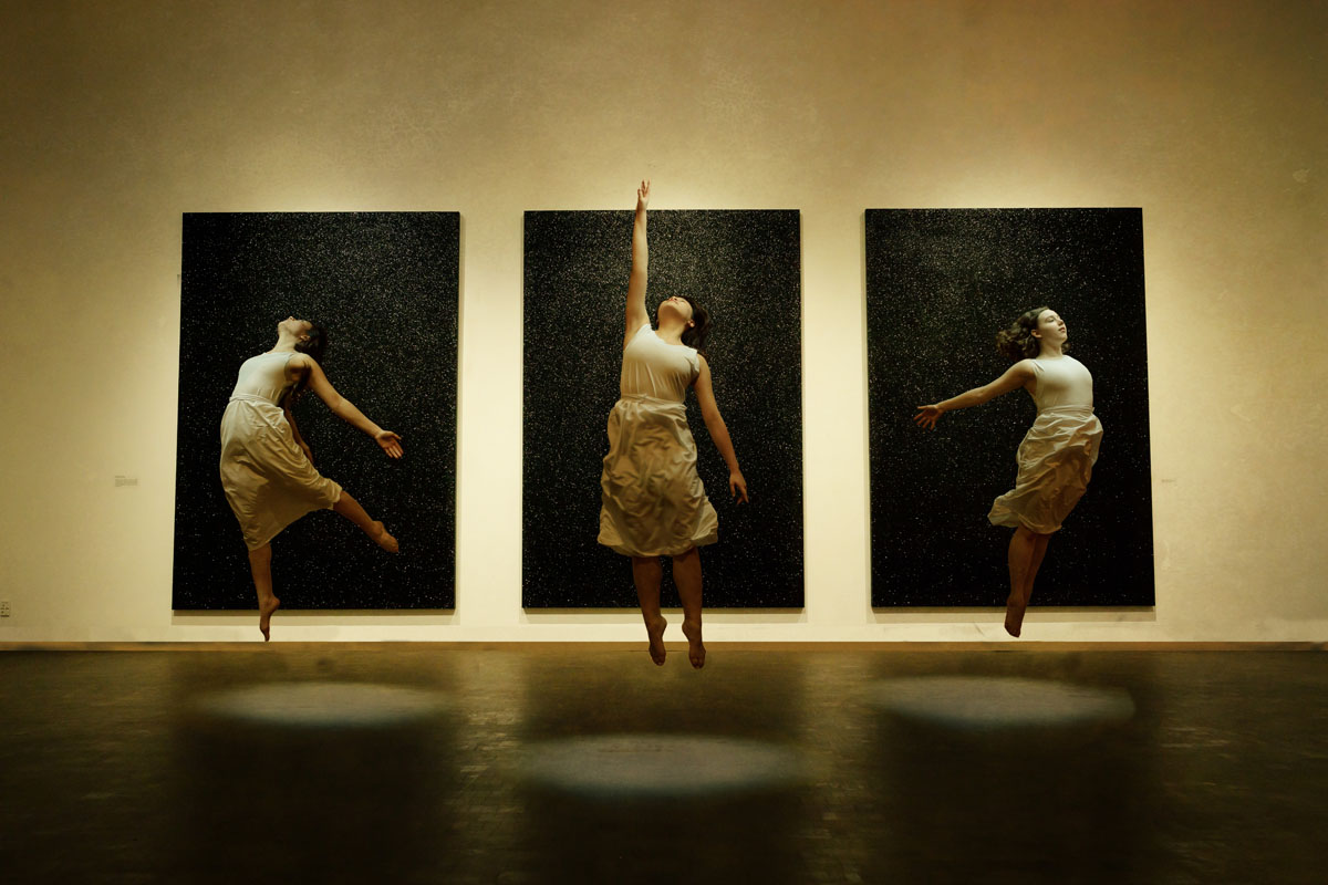 Dancers in the Western Gallery hover in the air in white dresses against a three black paintings.