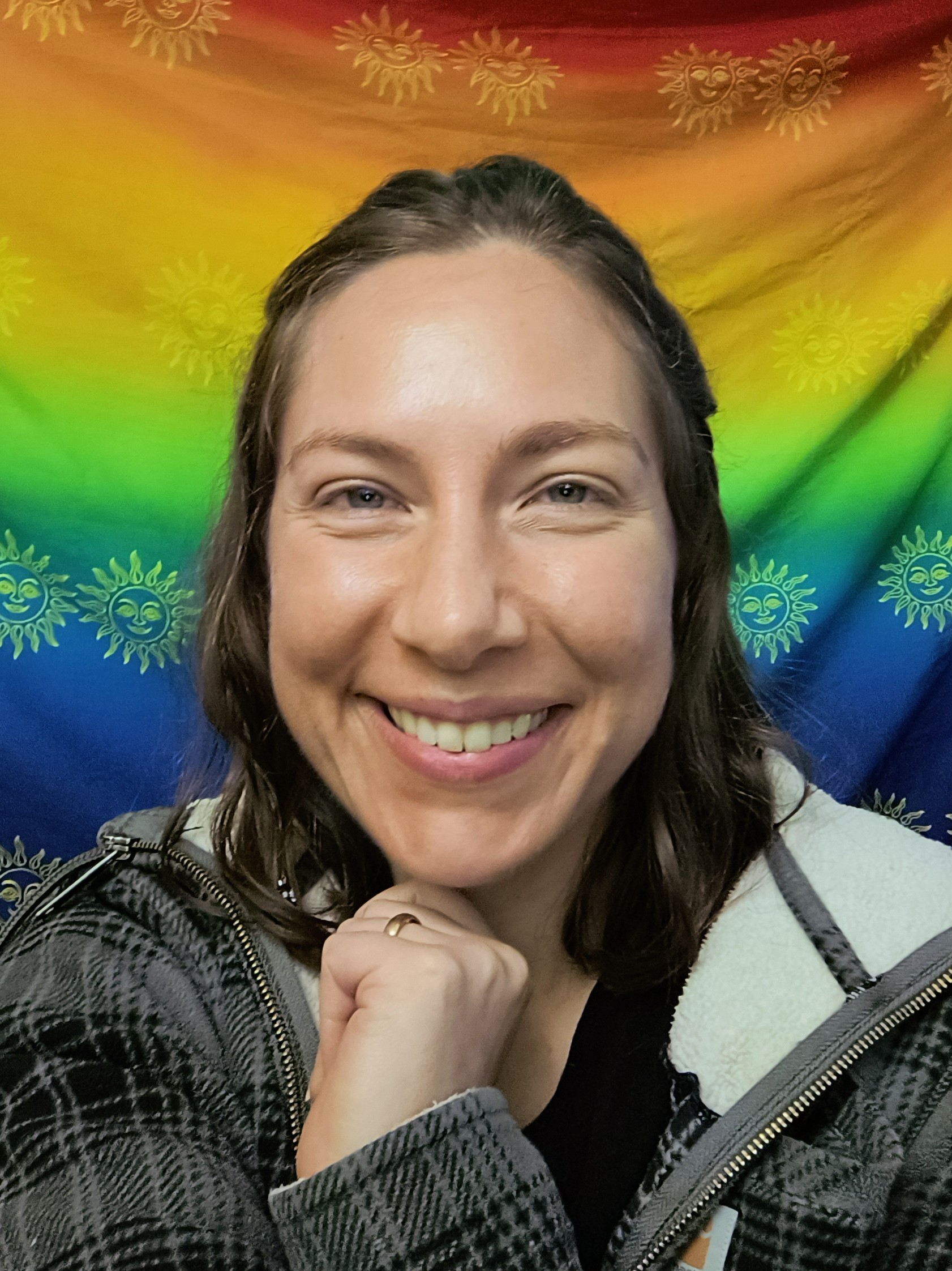 Justene wears a plaid fleece jacket and shoulder-length hair. She smiles in front of a rainbow cloth decorated with suns.