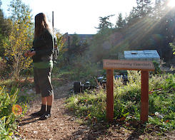 An individual wearing outdoor clothing standing near a handmade wooden sign that reads community garden