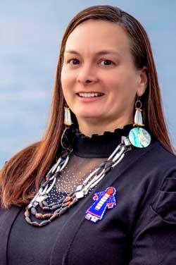 Theresa stands in front of rippling water, native beaded necklaces draped around her neck and large beaded earrings decorating her ear lobes.