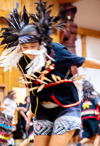 A young dancer dancing while wearing a head dress with black and white feathers and a wooden necklace.