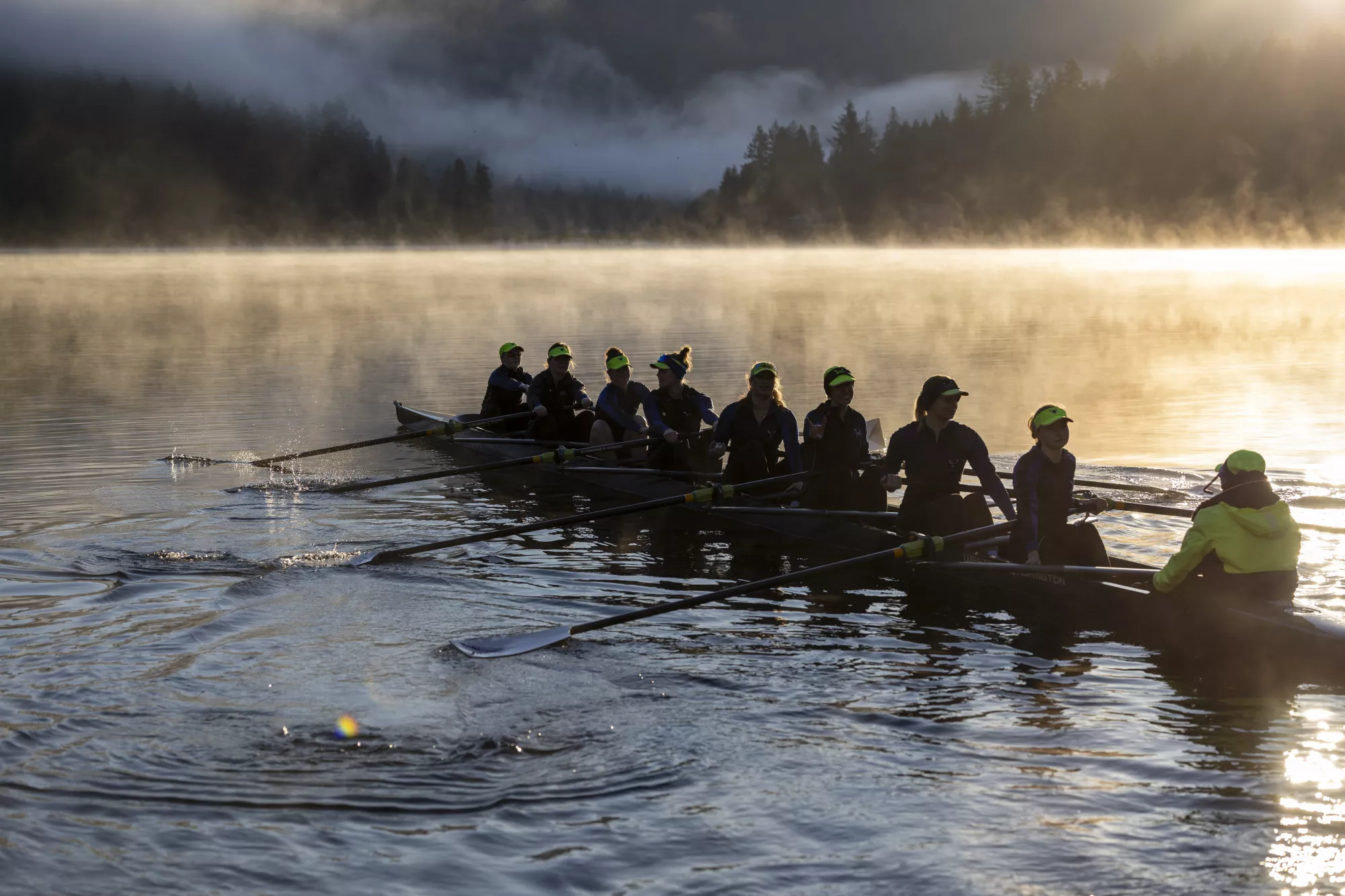 Members of the WWU rowing team row a boat across a misty lake with trees on the horizon.