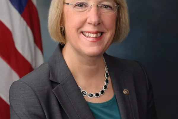 Senator Patty Murray wears a gray jacket and stands in front of the American flag.
