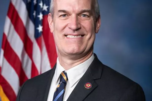 Rick Larsen in a button down collared shirt and tie, stands in front of an American flag.