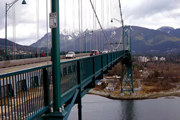 Against a background of mountains, cars cross a suspension bridge that takes them to the city of Vancouver BC.