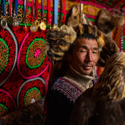 A Mongolian man standing in a bright yurt with a golden eagle on his arm. He wears a fur hat. Medals are hung on the richly embroidered walls of the yurt.