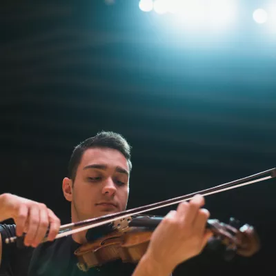 A student playing a violin on a stage under lights.