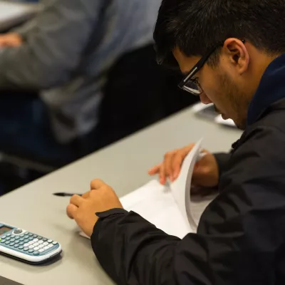 A student sitting at a table looking over papers with a calculator next to him.