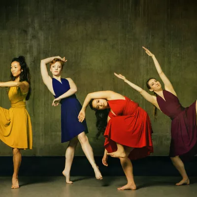 4 dancers in various poses wearing yellow, navy, red, and plum dresses.