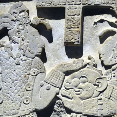 Mayan stone carving of a royal lady giving her king a jaguar mask and shield. They wear elaborate clothing, headdresses, and body jewelry.