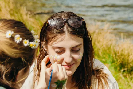 A student sitting on the lake shore on a sunny day having their face painted, a white flower motif