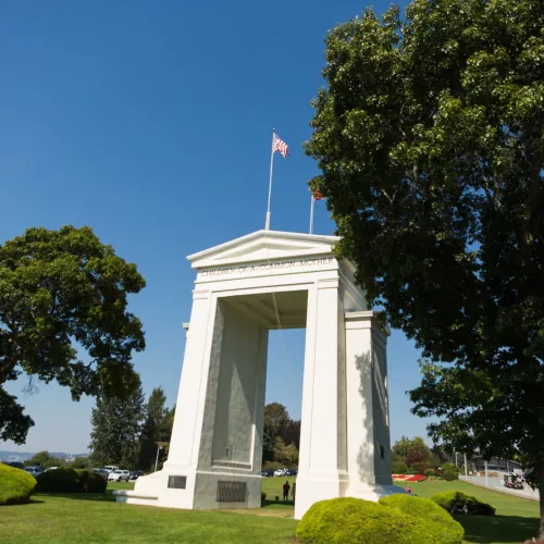 A large white archway sits on a lawn surrounded by trees, with US and Canadian flags flying on the top of the arch.