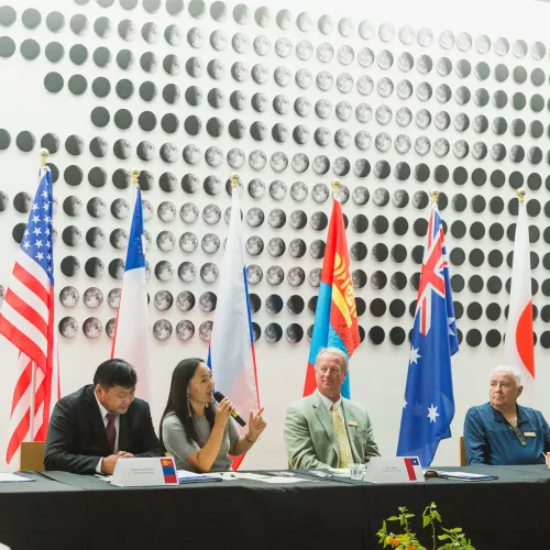 A panel of six people sit at a table, with many international flags displayed behind them.