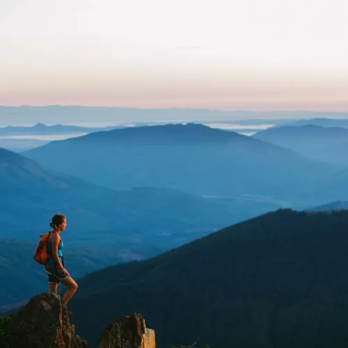 A hiker stands on a mountain peak overlooking a hilly landscape.