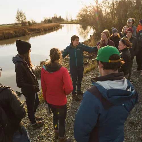 An instructor and group of students stand at the banks of a waterway.