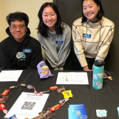 Three students standing at a table with give away items on it tabling for an event