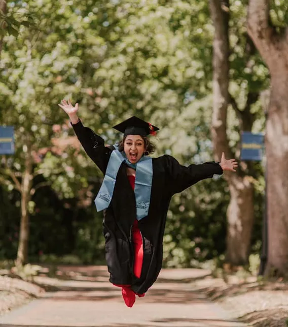 Amrit Abbasi jumping for joy in a graduation cap and gown with light blue sash