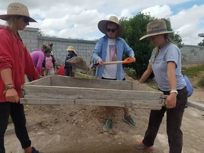 2 students lifting a wooden garden bed off the ground as another student shovels dirt into it.