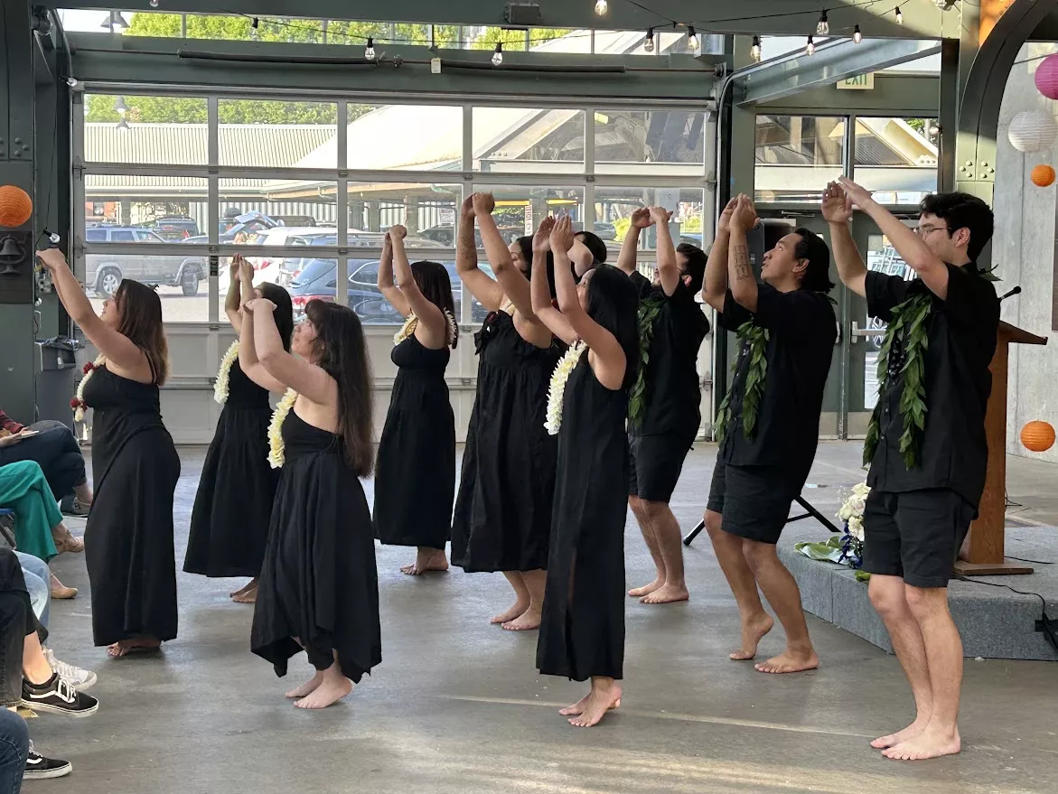 A group of people wearing black outfits and white/green leis, doing a traditional Polynesian dance