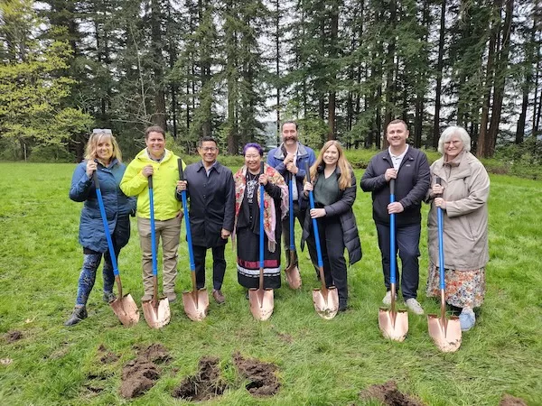 8 individuals standing with gold colored shovels outside on grass in front of a stand of evergreen trees.