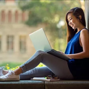 Woman sitting in a window overlooking the campus, working online with a laptop with her back against the sill. 