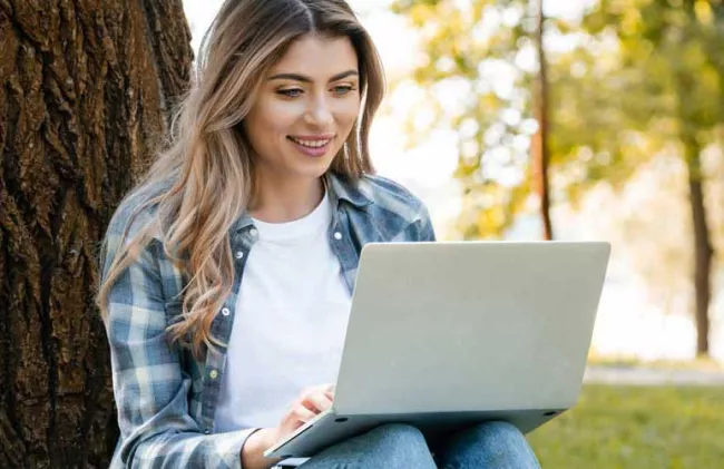 A female student sits quietly working on her online courses with her laptop. She is outside on a grassy, well landscaped lawn with her back against the rough bark of tree.
