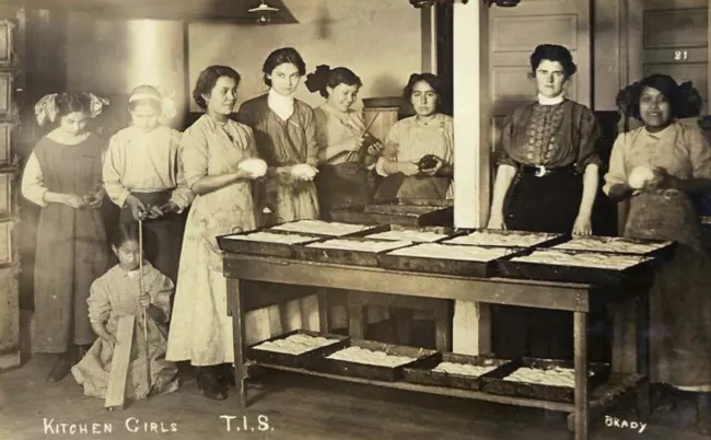 A group of Native American women and children work in the kitchen in front of a table lined with bread pans full of dough.