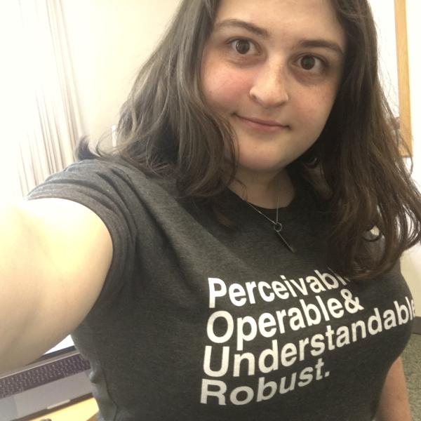 Carly wears a shirt with the text Perceivable and Operable and Understandable and Robust. She has light skin, shoulder-length brown hair and big eyes. 
